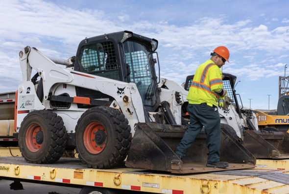 A white bulldozer loaded on top of a flatbed truck with a construction worker. 